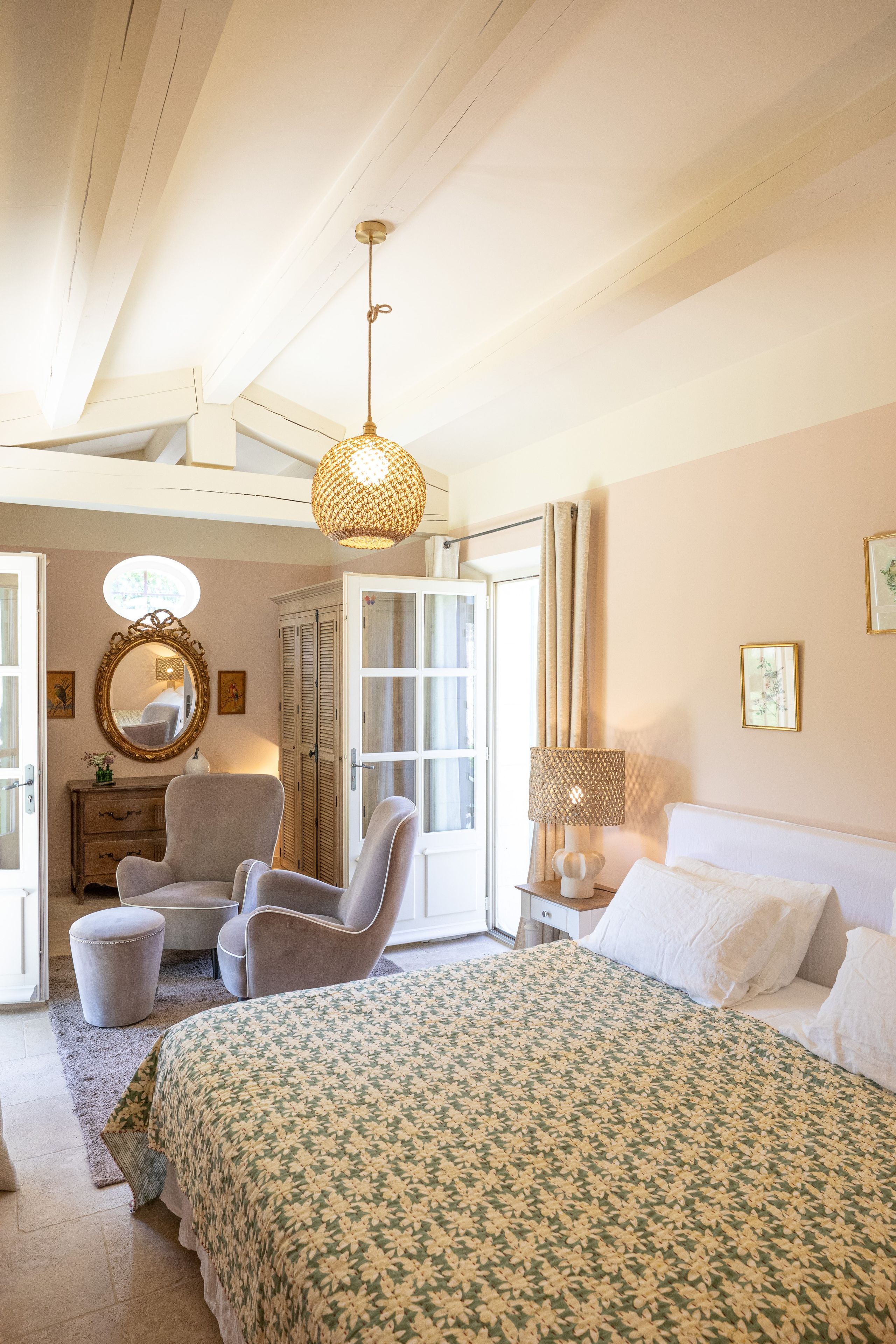 A bedroom with a bed, two chairs, a dresser, a mirror, a hanging light fixture, and a window.