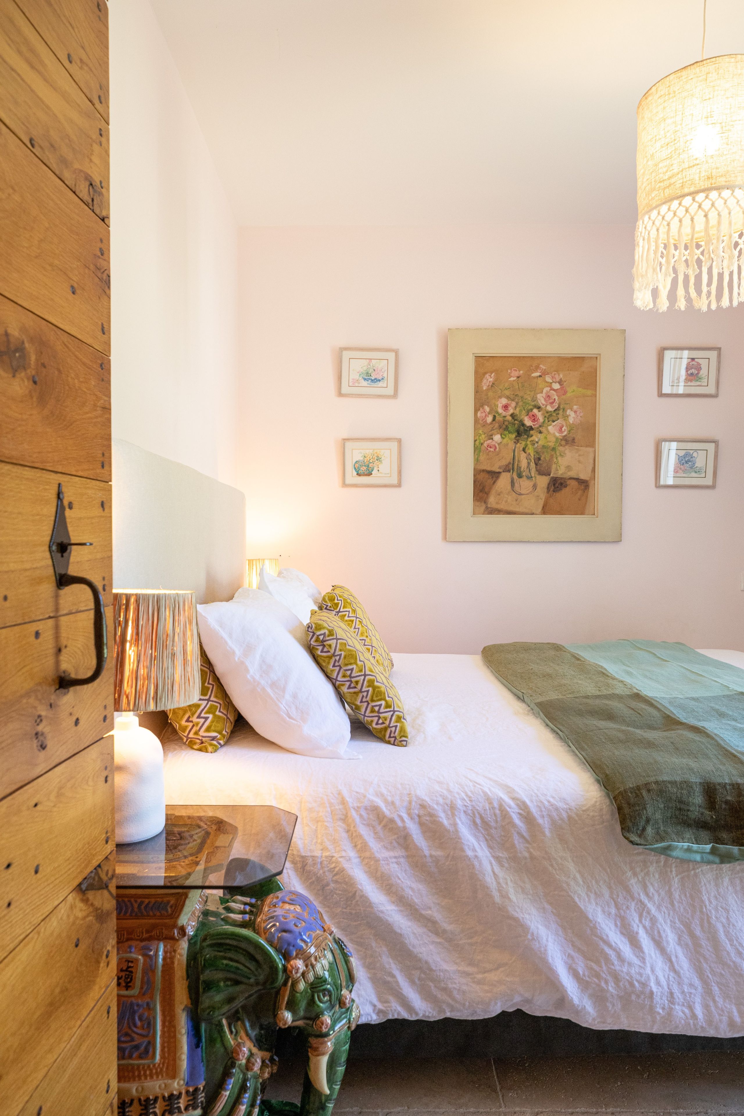 A bedroom with a bed, pillows, a bedside table with a lamp, a hanging light fixture, and a wall with framed pictures.