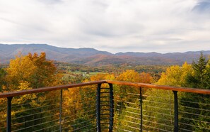 Ein malerischer Ausblick von einem Balkon mit Metallgeländer auf ein Tal mit herbstlich gefärbten Bäumen und entfernte Berge.