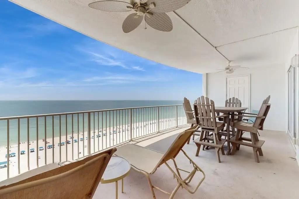 A balcony with a view of the beach, a table and chairs, and a ceiling fan.