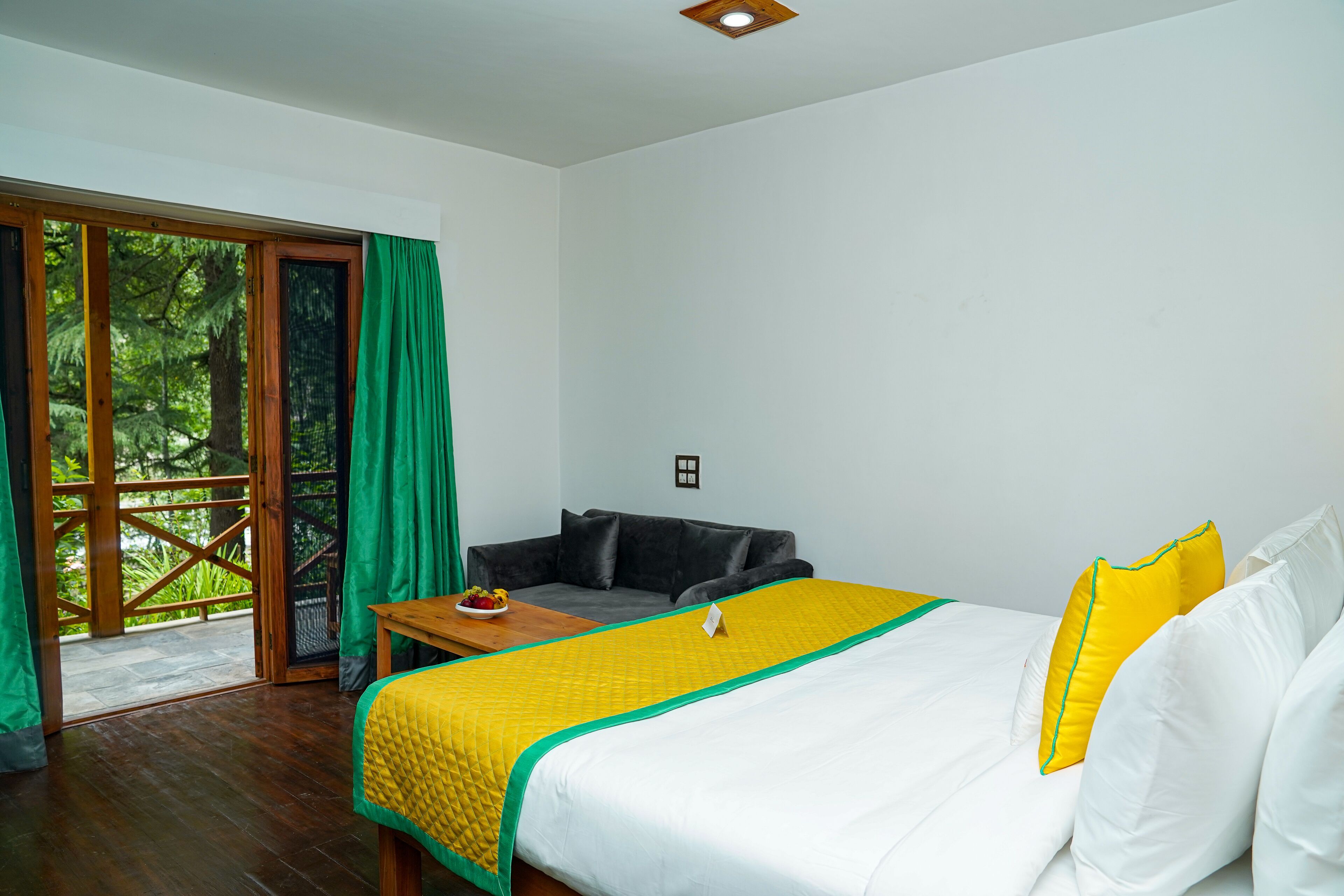 A hotel room with a bed, a sofa, a table with fruit, and a view of greenery through a sliding door.