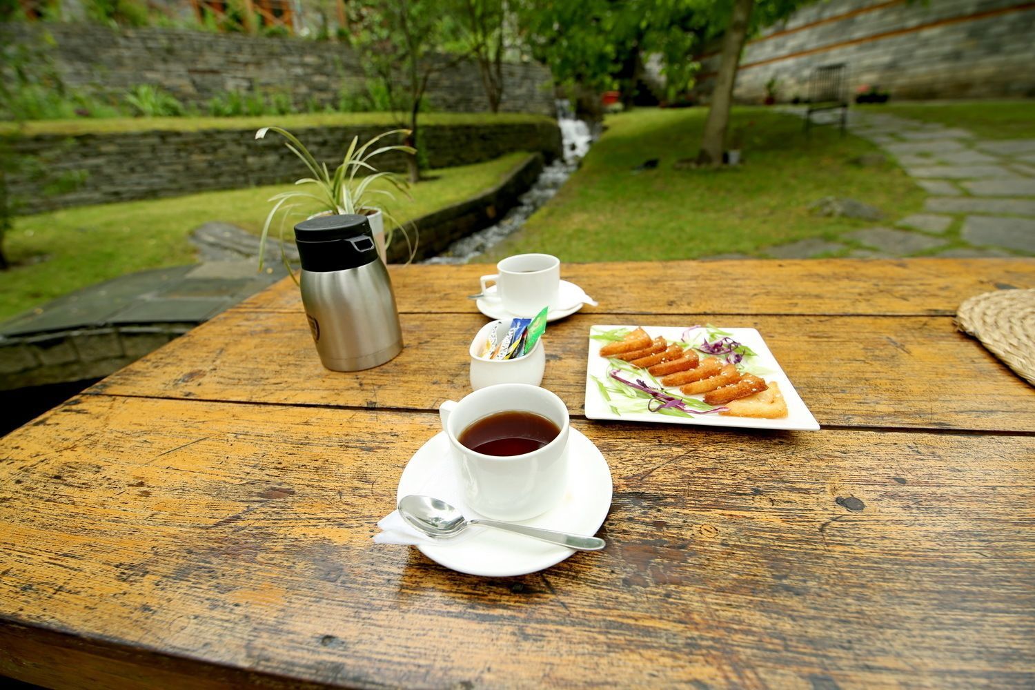 A wooden table with a cup of coffee, a thermos, and a plate of food.