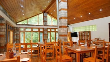 A dining area with wooden tables and chairs, stone walls, and large windows allowing natural light.