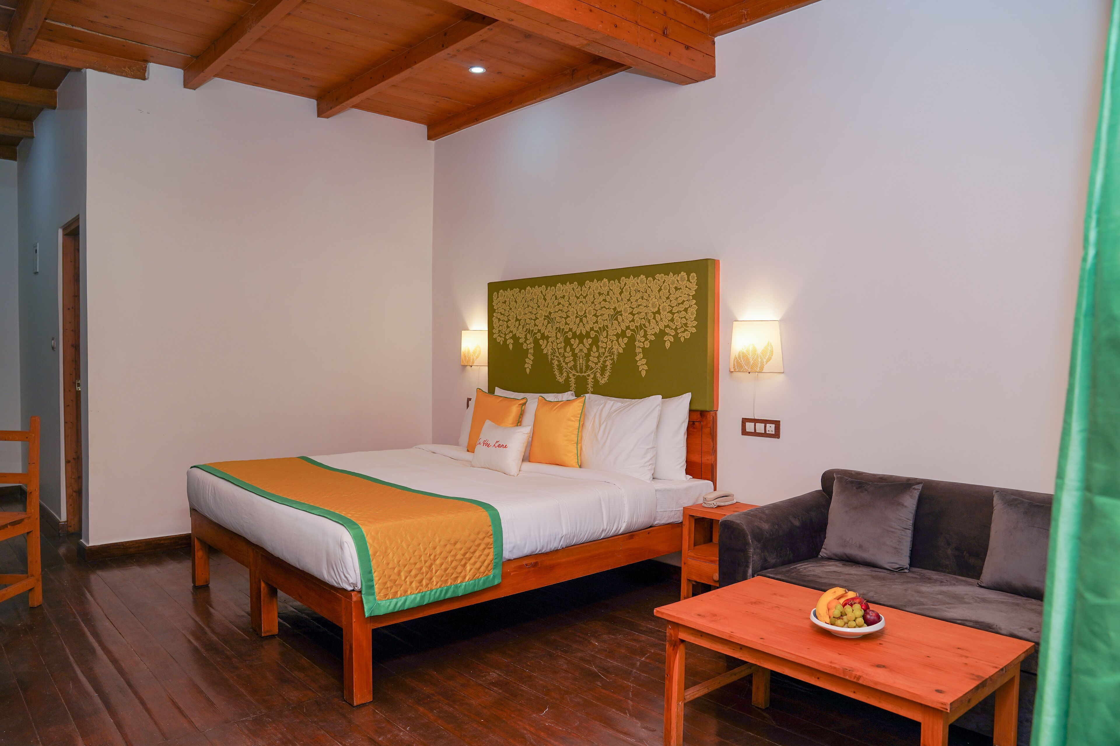 A hotel room with a wooden bed, a green headboard, a bedside table with a fruit bowl, and a grey sofa.