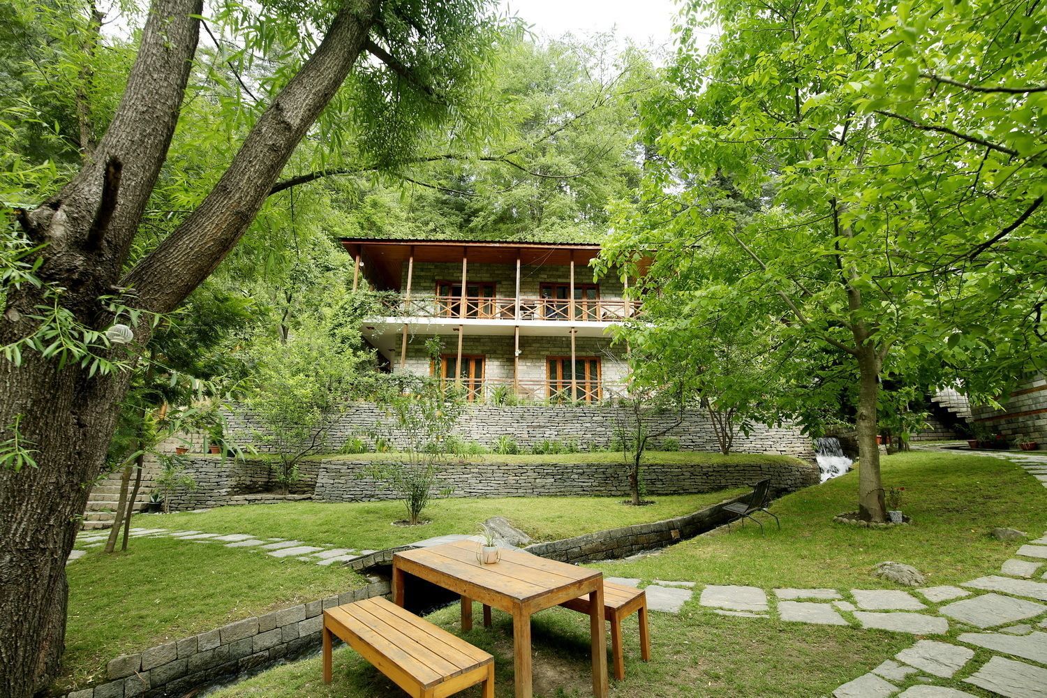 A two-story stone building with a balcony, surrounded by a well-maintained garden with a stone wall, a wooden table and bench, and a small waterfall feature.