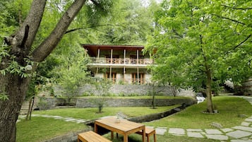 A two-story stone building with a balcony, surrounded by a well-maintained garden with a stone wall, a wooden table and bench, and a small waterfall feature.