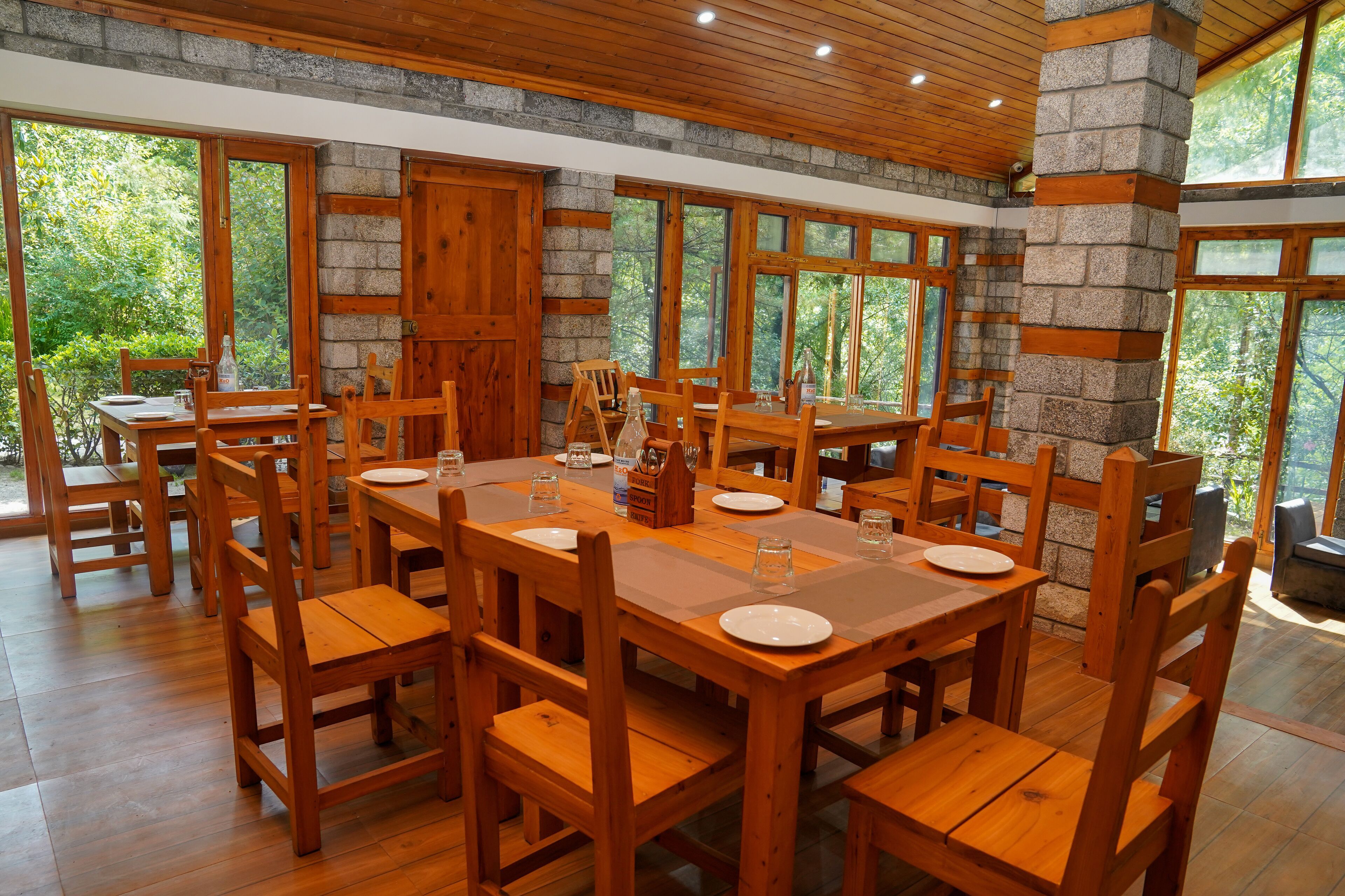 A dining area with wooden tables and chairs, stone walls, and large windows overlooking greenery.