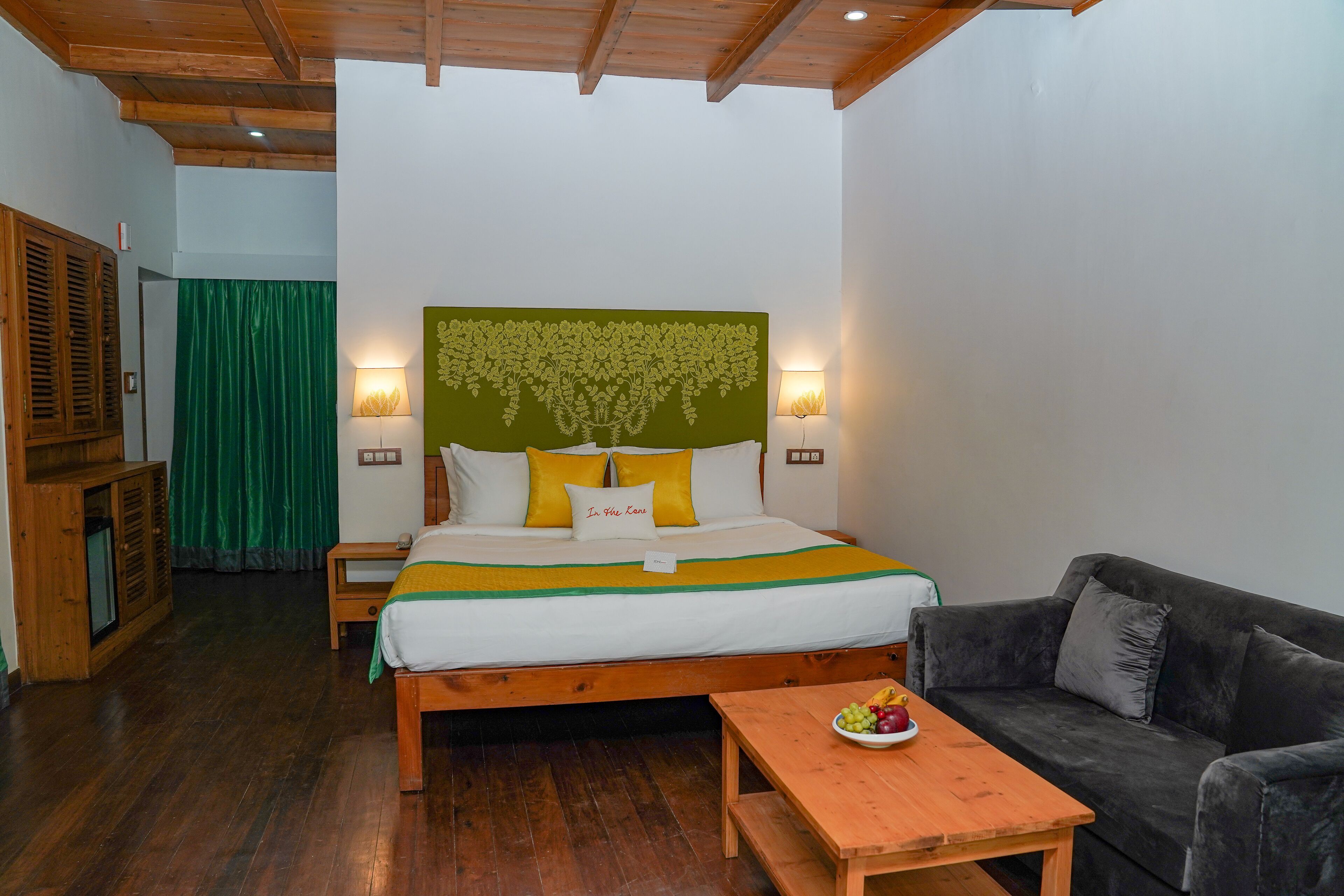 A hotel room with a bed, a wooden headboard, a green wall decoration, a grey sofa, and a wooden table with fruit.