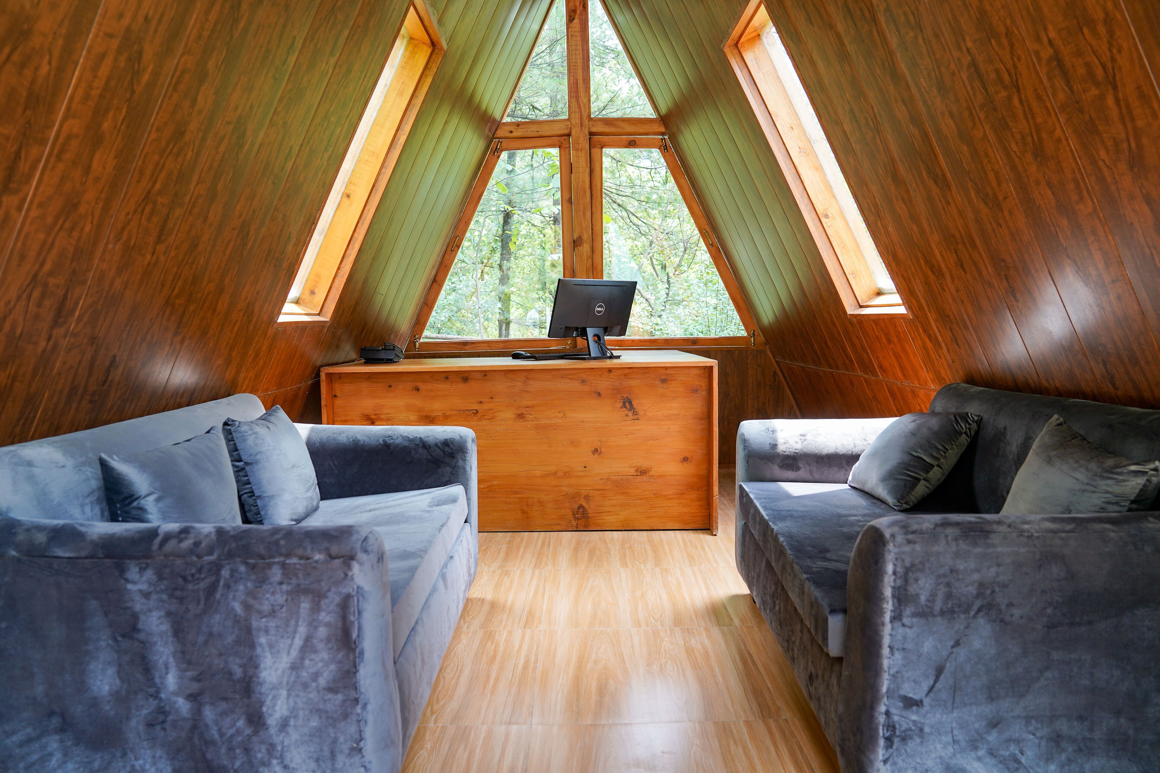 A cozy attic room with a wooden desk, two grey sofas, and a large window offering a view of trees.
