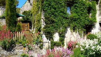 A stone building with a wooden staircase leading to a door, surrounded by lush greenery and flowering plants.