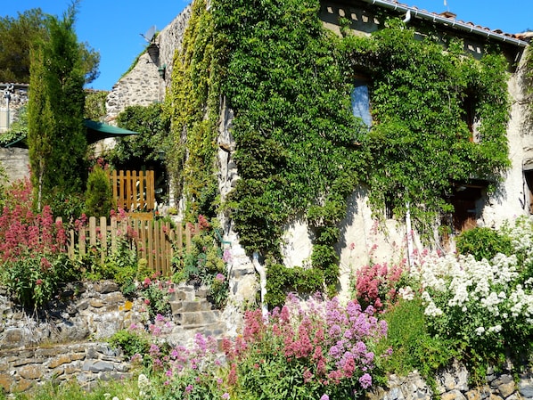 A stone building with a wooden staircase leading to a door, surrounded by lush greenery and flowering plants. - Holiday Home 'L'ecurie' with Mountain View (Vira)