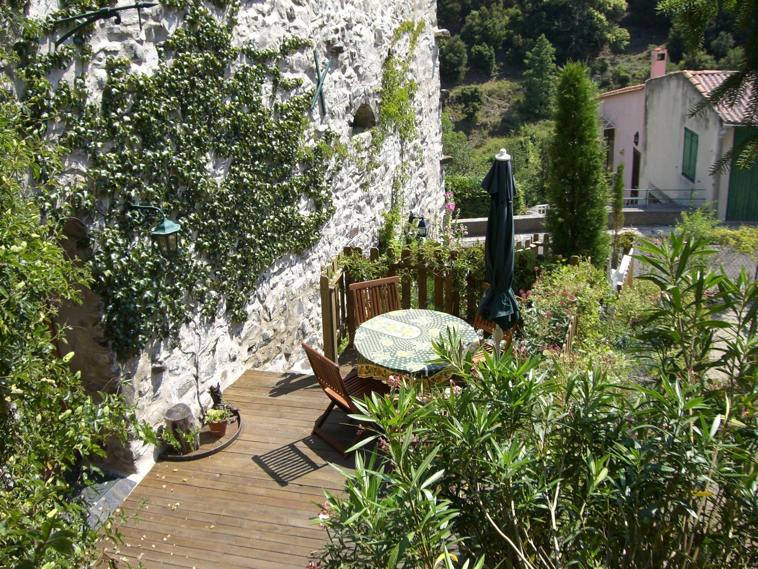 Une terrasse en bois avec une table et des chaises, entourée de verdure et d’un mur en pierre.