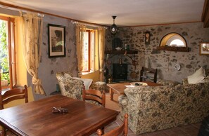 A rustic living room with a stone wall, a fireplace, a dining table, and floral-patterned furniture. - Holiday Home 'L'ecurie' with Mountain View (Vira)