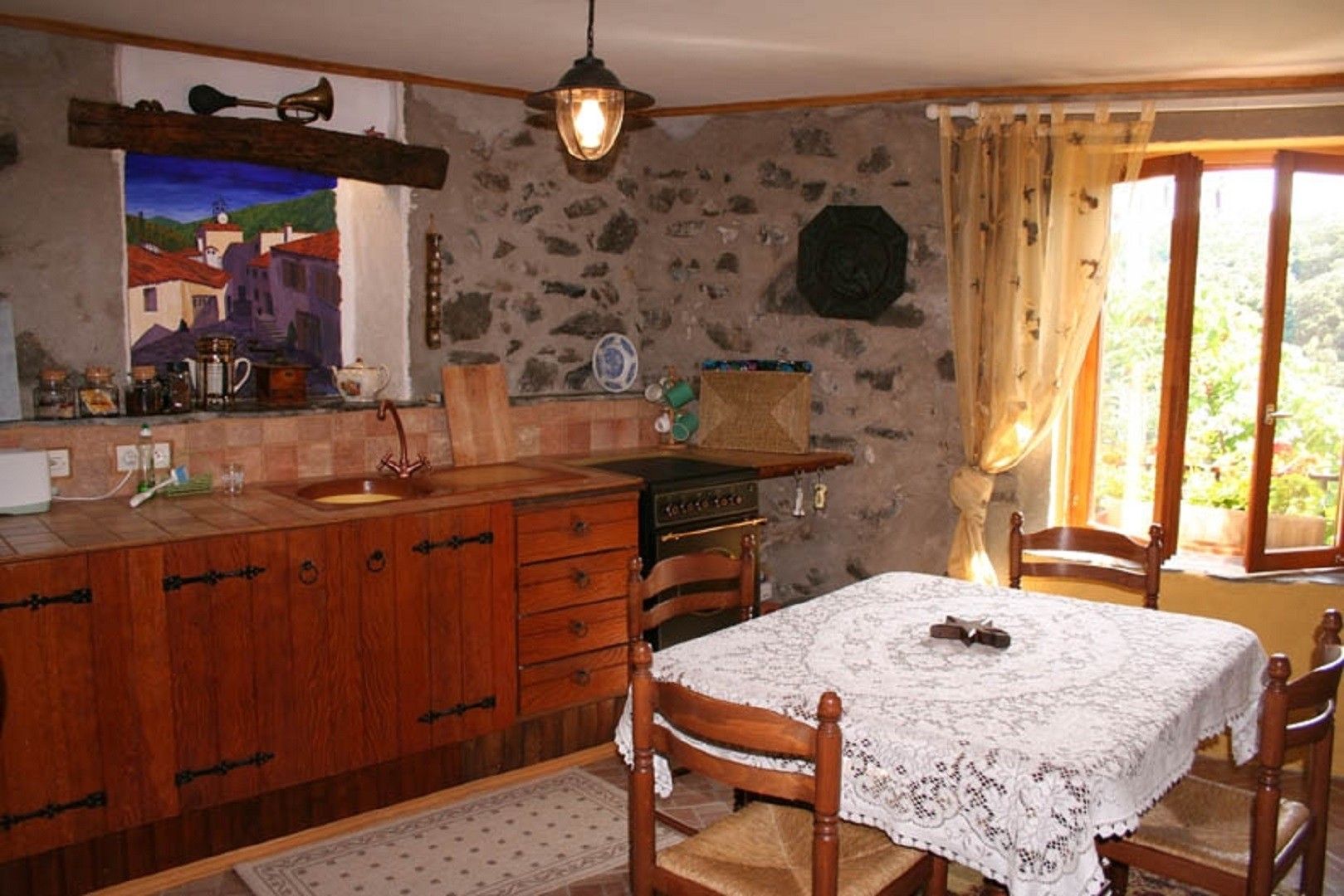 A rustic kitchen with a stone wall, wooden cabinets, a vintage stove, and a dining table with a lace tablecloth.