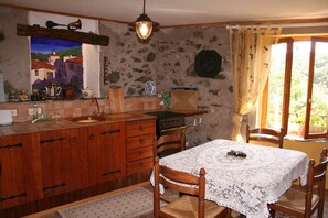 A rustic kitchen with a stone wall, wooden cabinets, a vintage stove, and a dining table with a lace tablecloth. - Holiday Home 'L'ecurie' with Mountain View (Vira)
