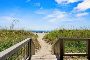 Ein Holzsteg führt zu einem Strand mit Sonnenschirmen und Liegestühlen.