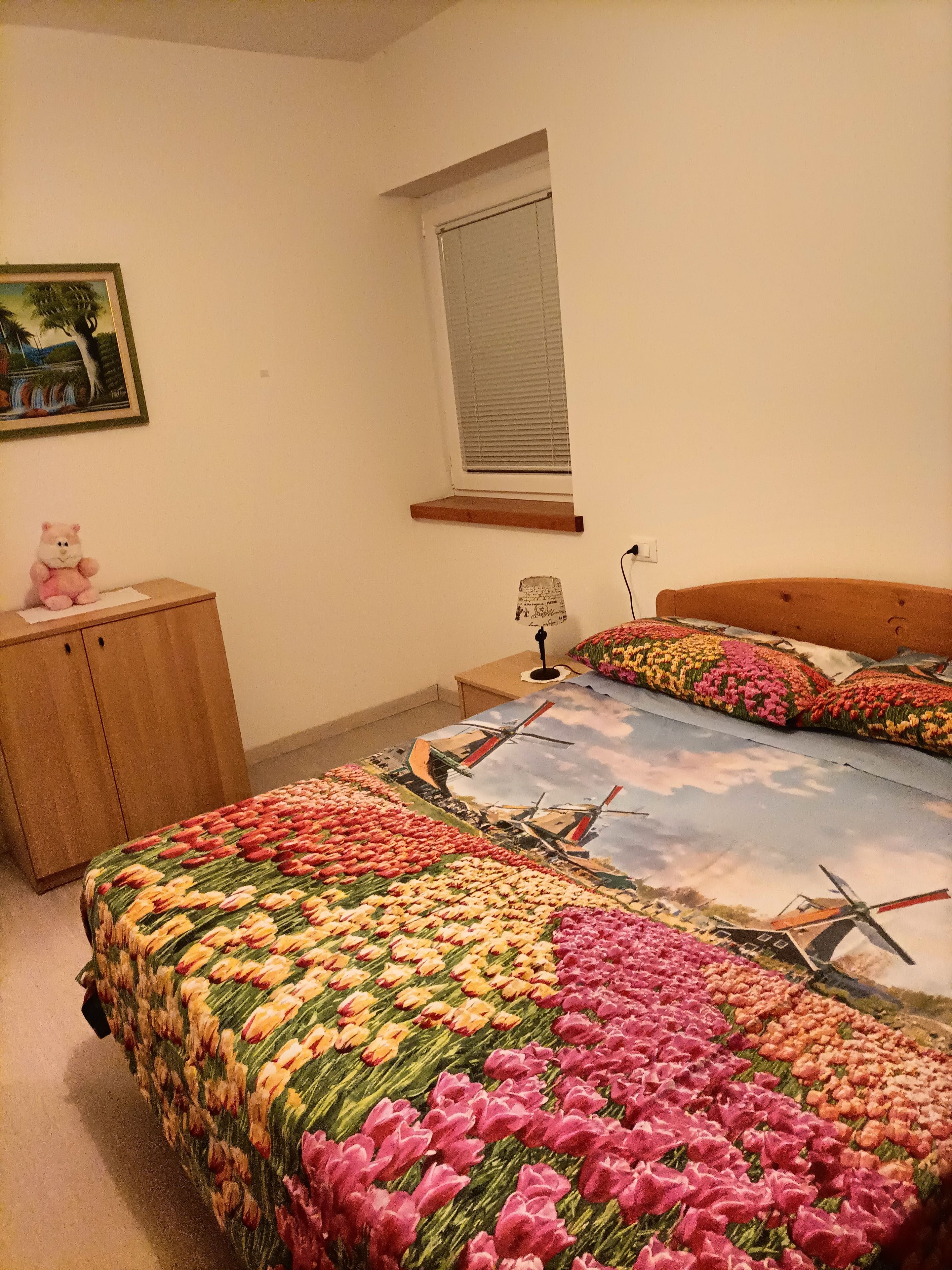 Ein Schlafzimmer mit einem Bett mit floralem und Windmüller-Motiv, einem Holznachttisch, einem kleinen Schreibtisch, einem Stuhl und einem Teddybär.