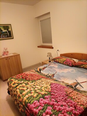 A bedroom with a bed featuring a floral and windmill design, a wooden nightstand, a small desk, a chair, and a teddy bear.