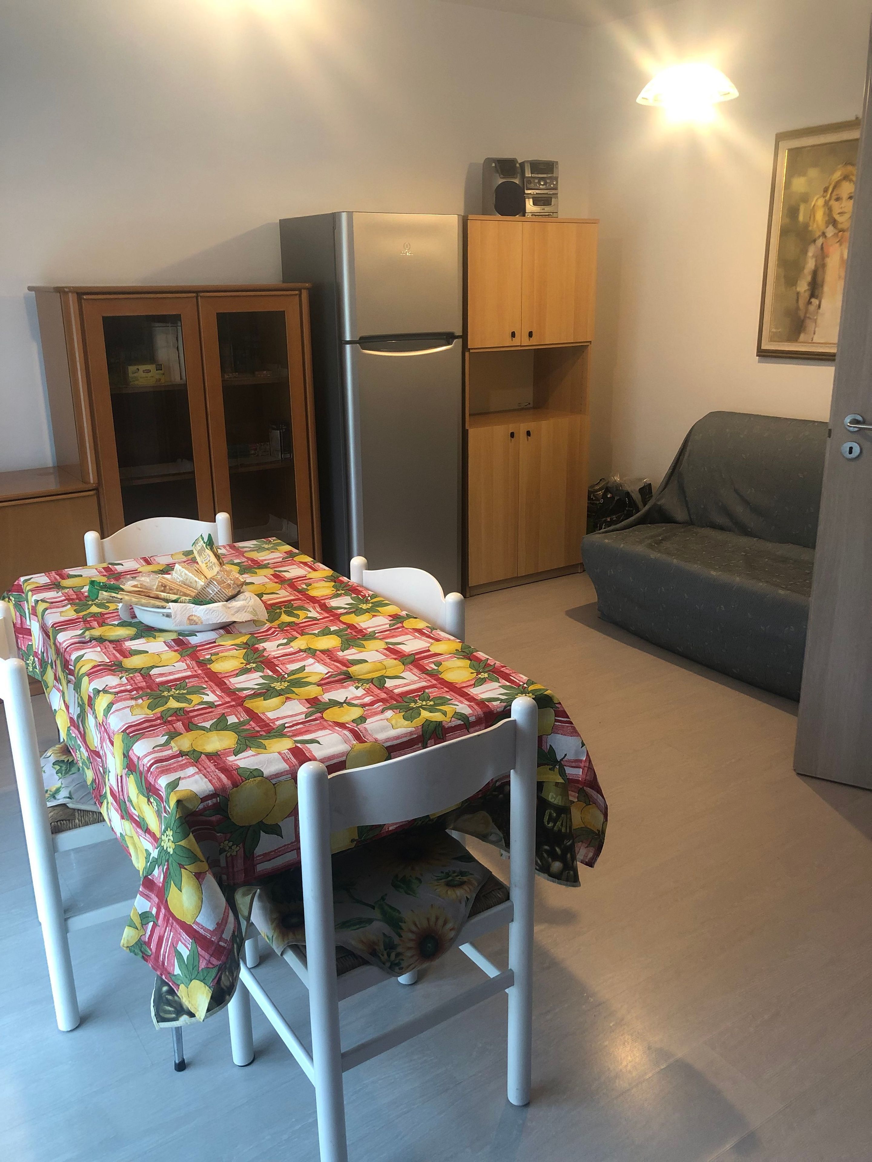 Eine kompakte Küche mit Kühlschrank, Holzschränken, einem Esstisch mit Obstmuster-Tischdecke und einem grauen Sofa.