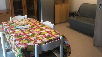Eine kompakte Küche mit Kühlschrank, Holzschränken, einem Esstisch mit Obstmuster-Tischdecke und einem grauen Sofa.