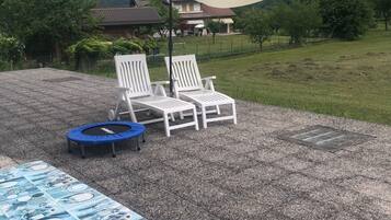 Eine Terrasse mit zwei weißen Liegestühlen, einem blauen Trampolin und einem Tisch mit blauer Tischdecke.