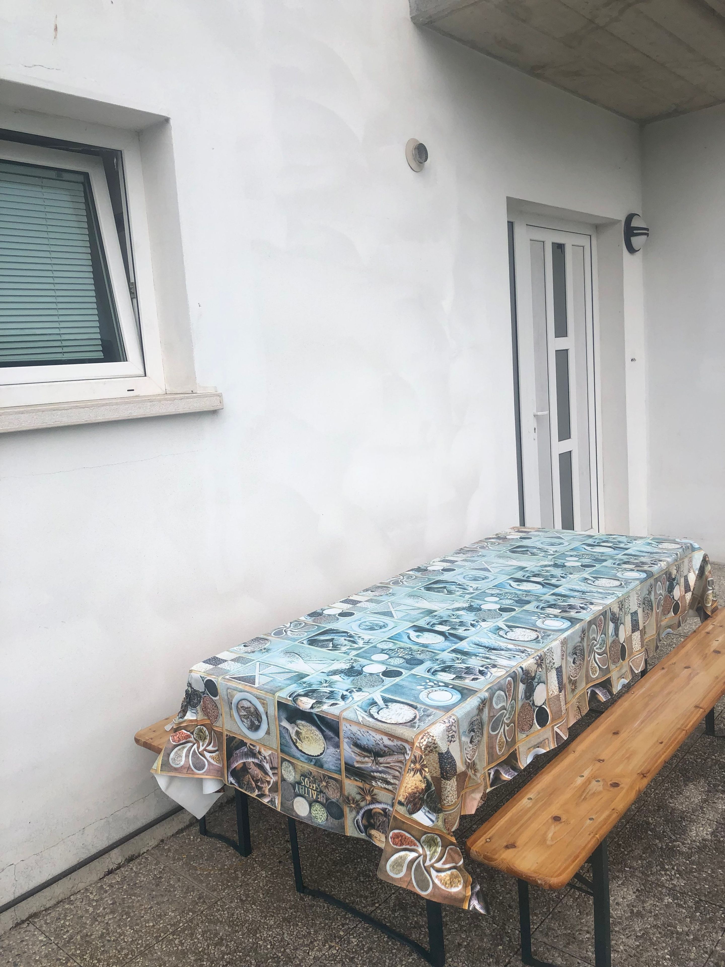 Ein Tisch im Freien, bedeckt mit einer Tischdecke mit Muster, eine Bank und eine weiße Tür.