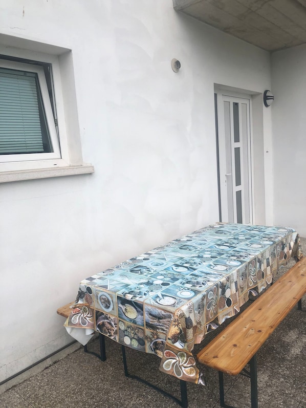 A covered outdoor table with a patterned tablecloth, a bench, and a white door.