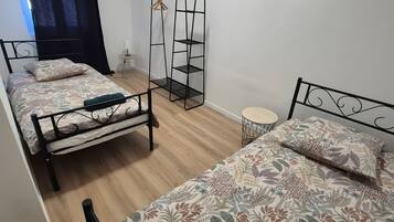 Ein Zimmer mit drei Einzelbetten, einem stehenden Kleiderschrank, einem kleinen runden Tisch und einem an der Wand befestigten Regal.