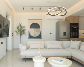 Un salon moderne comprenant un canapé d’angle, une table basse, une plante en pot, une œuvre d’art accrochée au mur et un coin cuisine équipé d’appareils électroménagers.