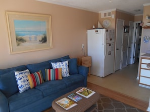 A living room with a blue sofa, a coffee table with magazines, a kitchen area with a refrigerator, and a painting on the wall. - A Place at the Beach (Isle of Palms)