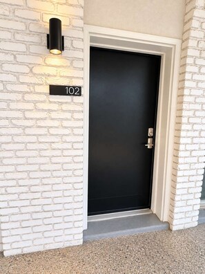 A black door with a number plate '102' on a white brick wall. - The Sunny Seaside 300 Tenth | 102 (Myrtle Beach)