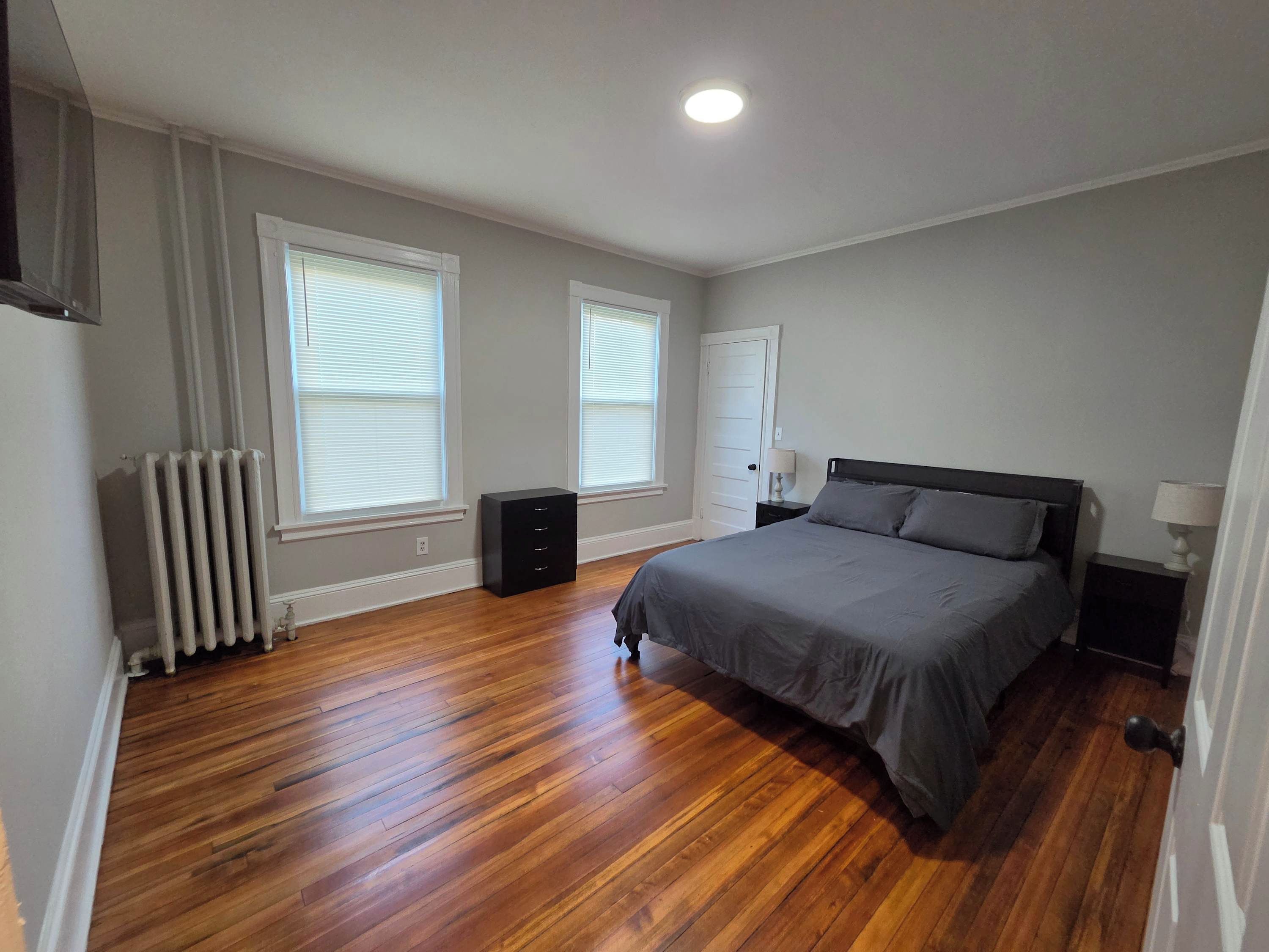 A bedroom with a bed, a radiator, a dresser, and two windows with blinds.