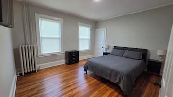 A bedroom with a bed, a radiator, a dresser, and two windows with blinds.