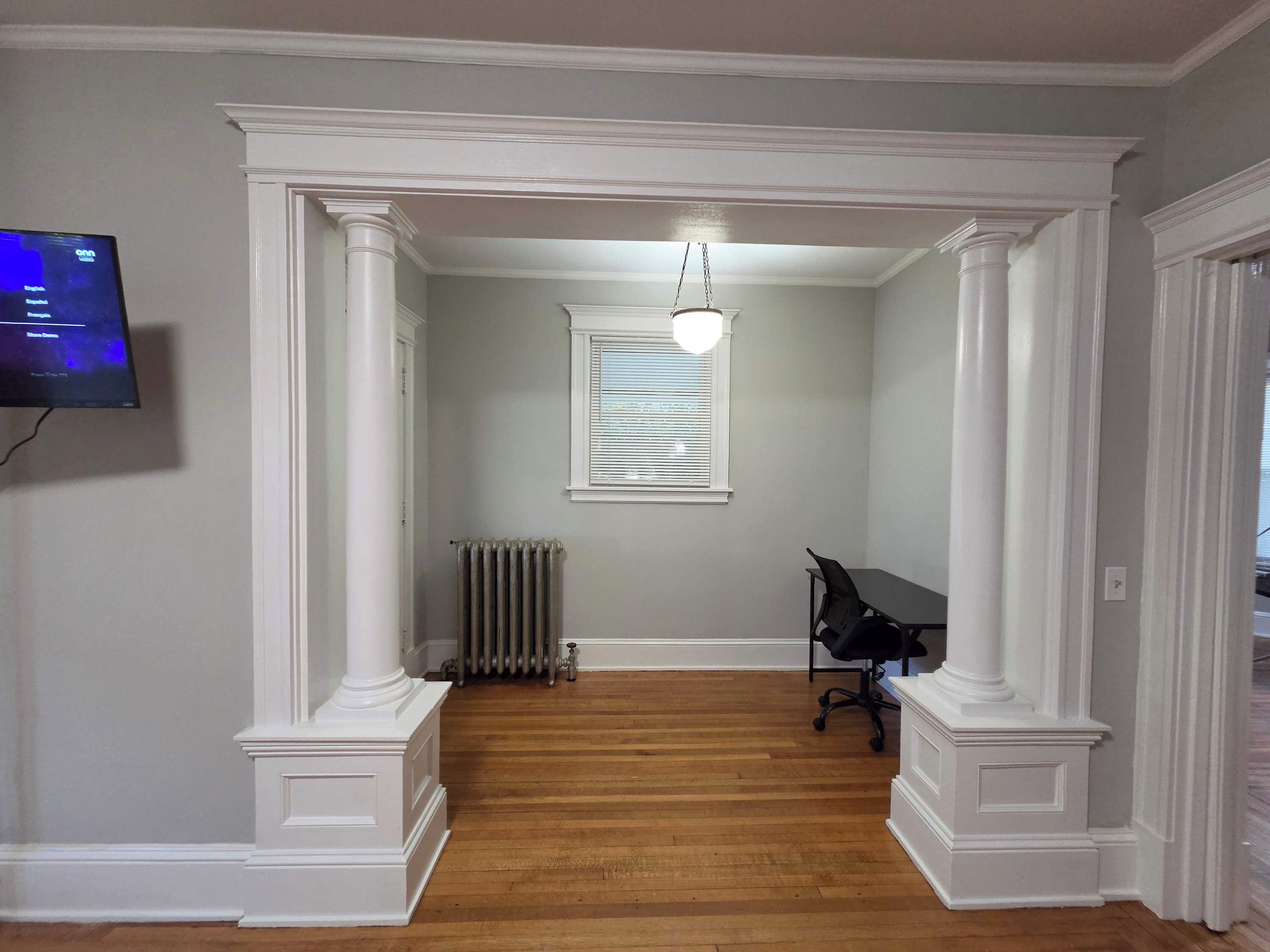 Un couloir avec un radiateur, un fauteuil de bureau noir et un téléviseur fixé au mur.