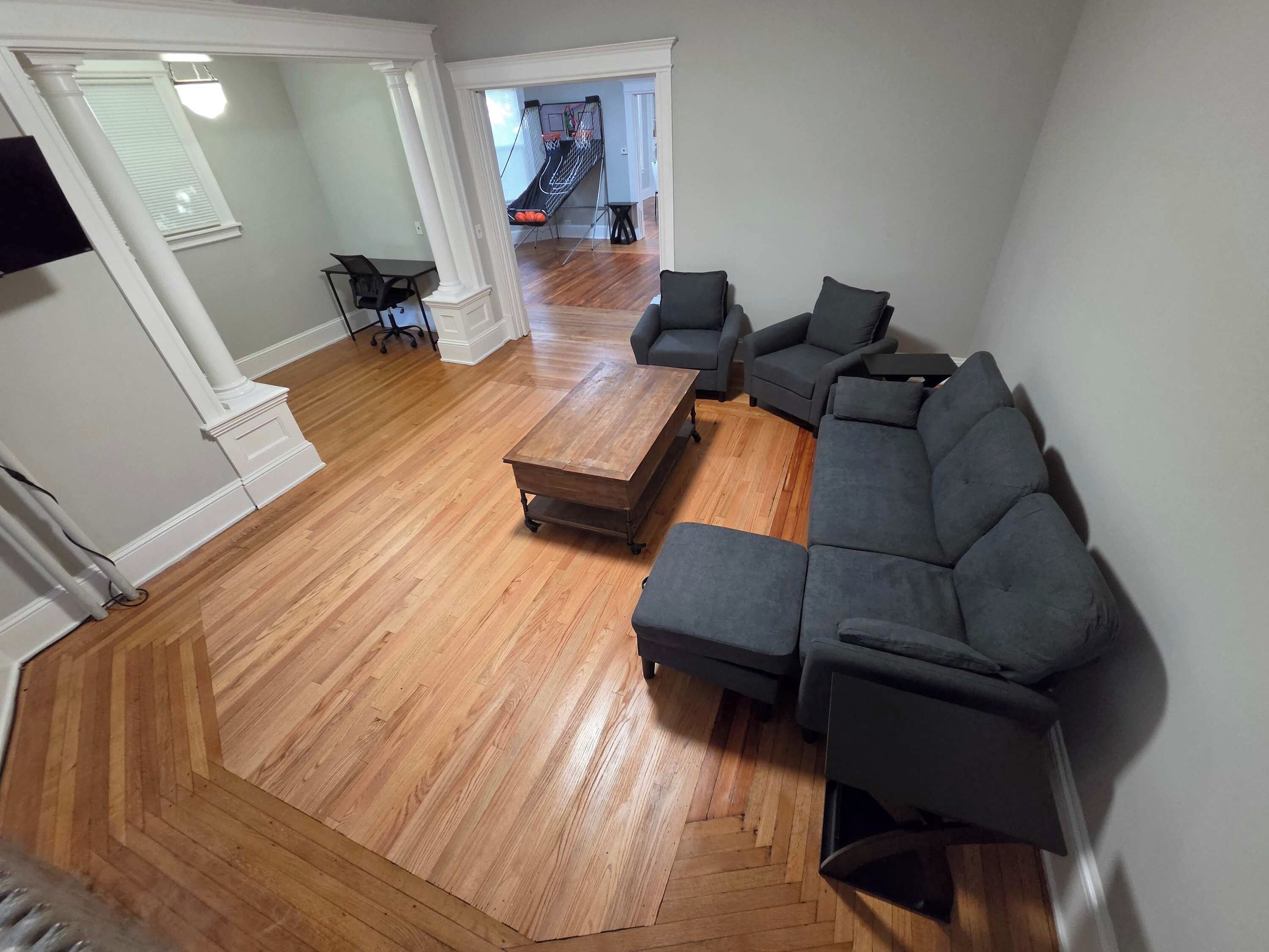 A spacious living room with a herringbone wood floor, a sectional sofa, a coffee table, and a small office chair.