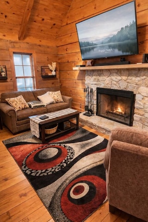 A cozy cabin interior with a stone fireplace, a television mounted above it, a wooden ceiling, and a large window. - Lakeview Cabin | Dock, Fire Pit & Screened Porch (Greencastle)