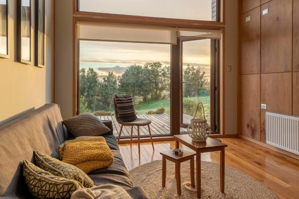 Una sala de estar moderna con un sofá, una silla, una mesita y vistas a una terraza exterior a través de un amplio ventanal corredero.