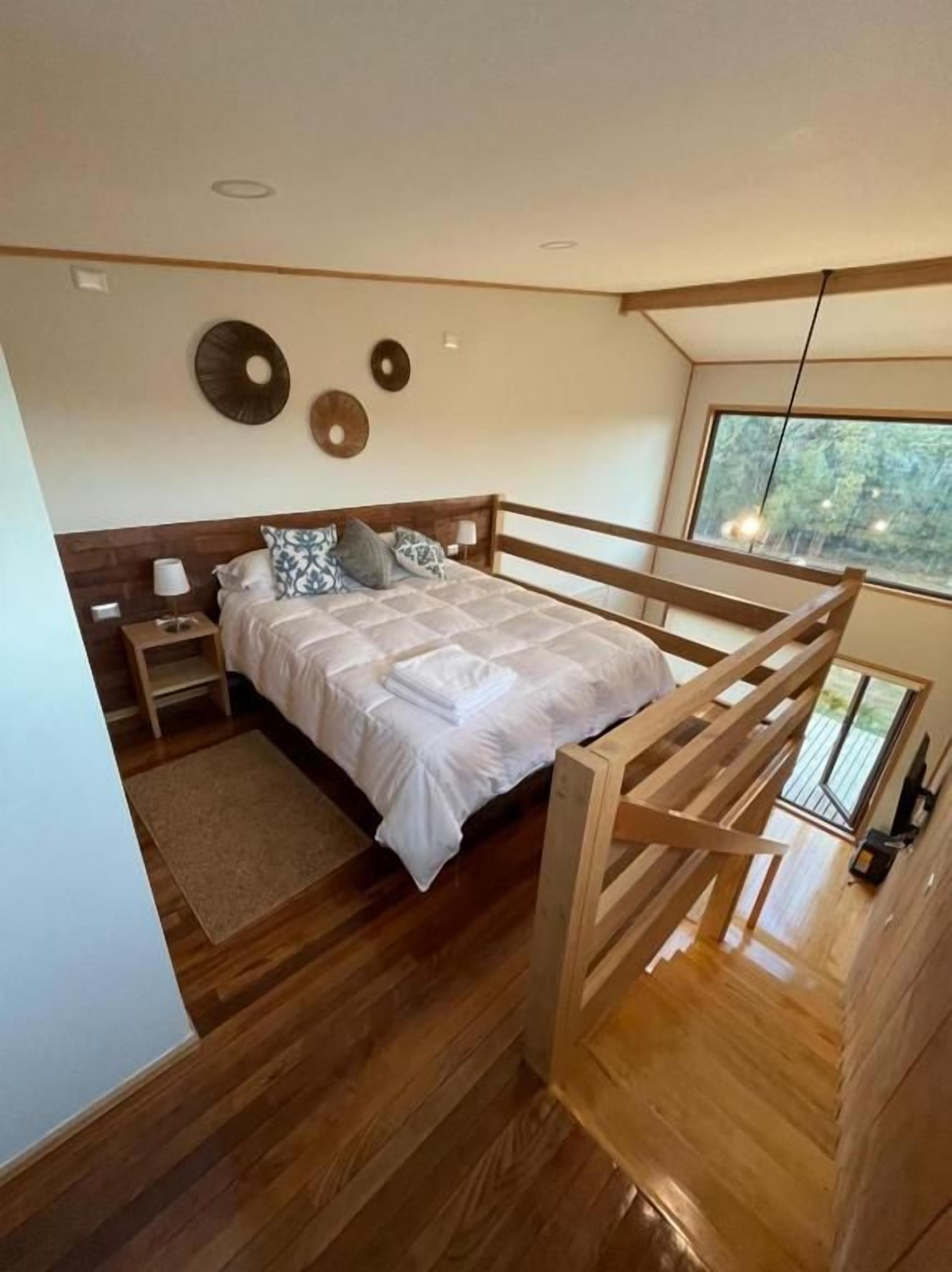 A modern bedroom with a wooden bed frame, a nightstand, and a bedside table. There is a window with a view of trees.