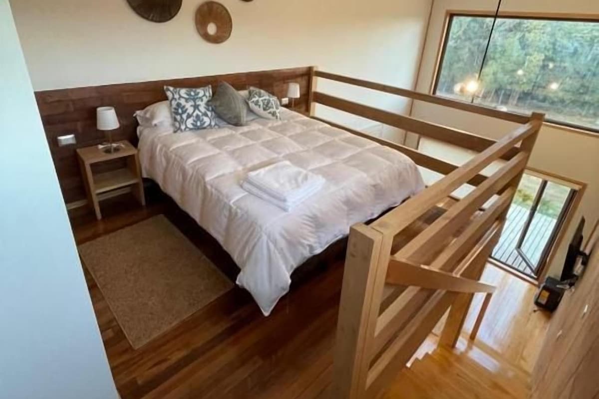 Ein modernes Schlafzimmer mit einem Bettgestell aus Holz, einem Nachttisch und einer Nachttruhe. Ein Fenster bietet Blick auf Bäume.