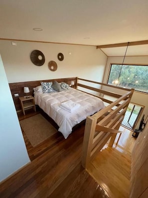 Ein modernes Schlafzimmer mit einem Bettgestell aus Holz, einem Nachttisch und einer Nachttruhe. Ein Fenster bietet Blick auf Bäume.