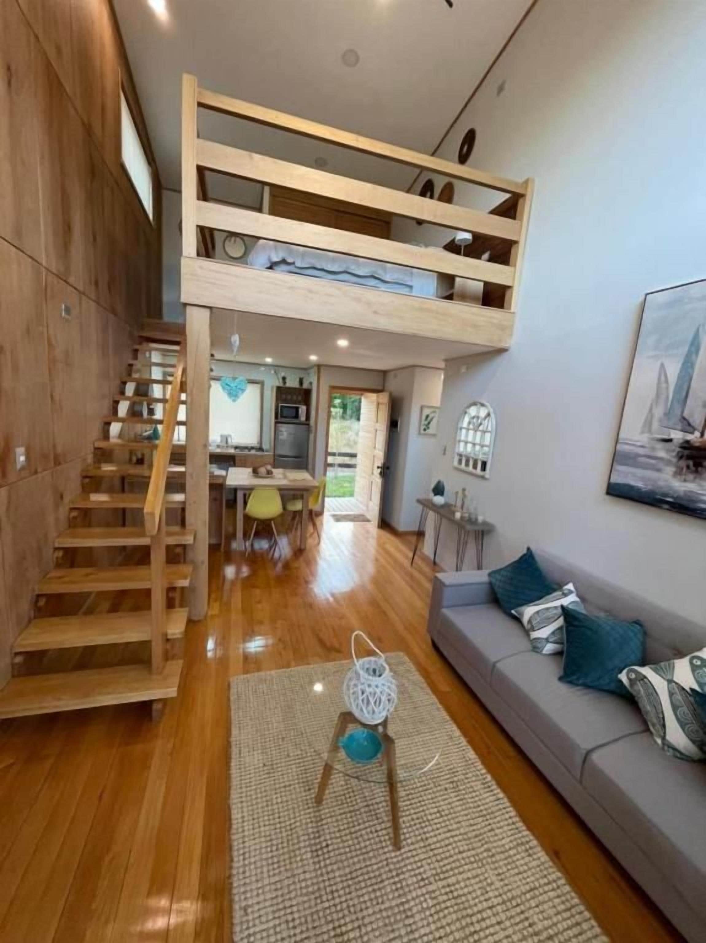 Un espacio tipo loft con una escalera de madera, un sofá con cojines y una zona de comedor al fondo.