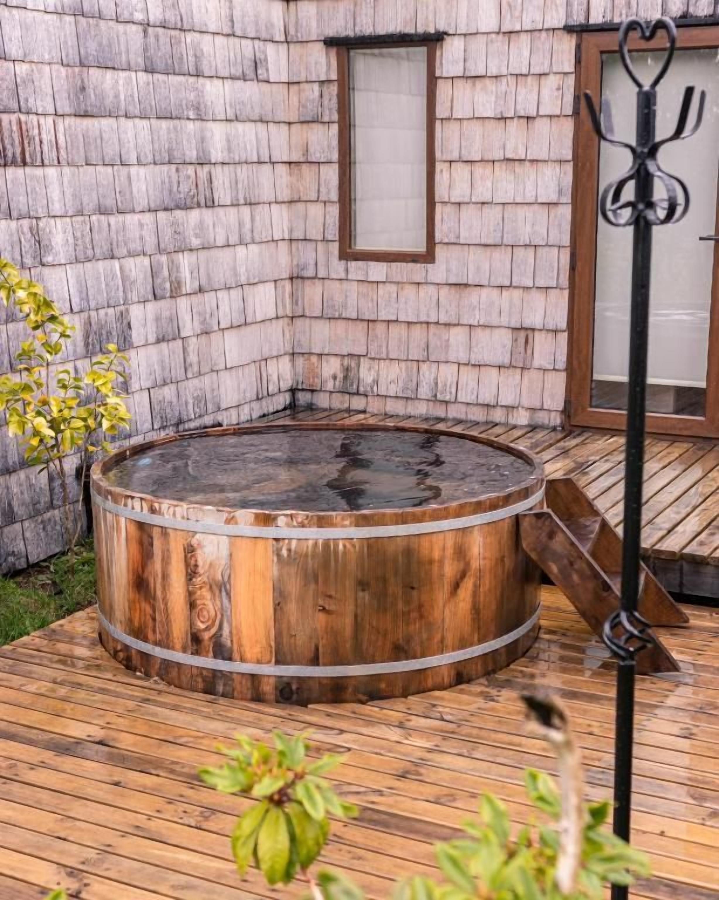 A wooden hot tub on a wooden deck with a brick wall and a window in the background.