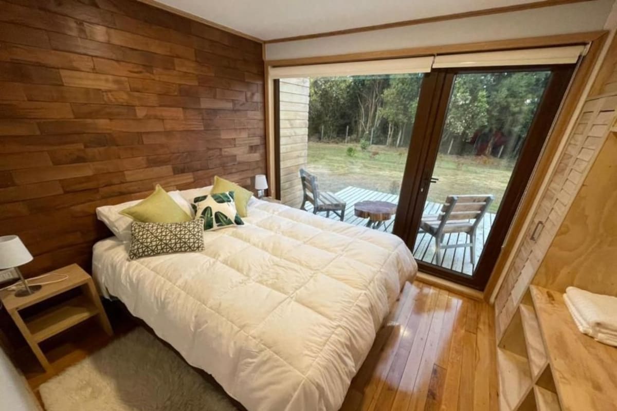 Ein Schlafzimmer mit einem großen Bett, einem Holzkopfteil, einem Nachttisch, einer Lampe und Blick auf eine Außenpatio mit Stühlen und einem Tisch.