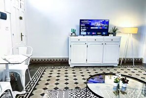A room with a television on a cabinet, a white chair, a small table with a glass top, and patterned floor tiles. - La Marsa Vibe (Tunis)