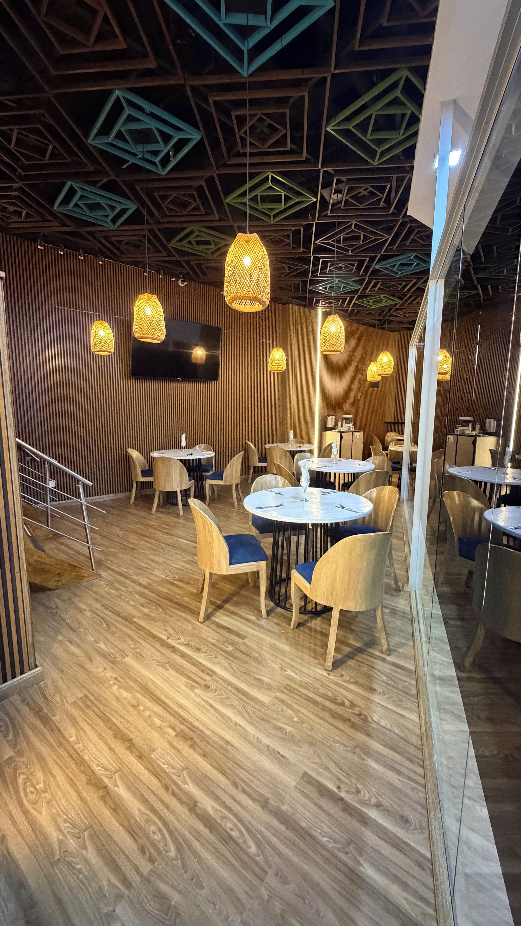 Een restaurantinterieur met houten vloeren, ronde tafels en stoelen, met geometrische patronen in het plafond en hangende pendantlampen.