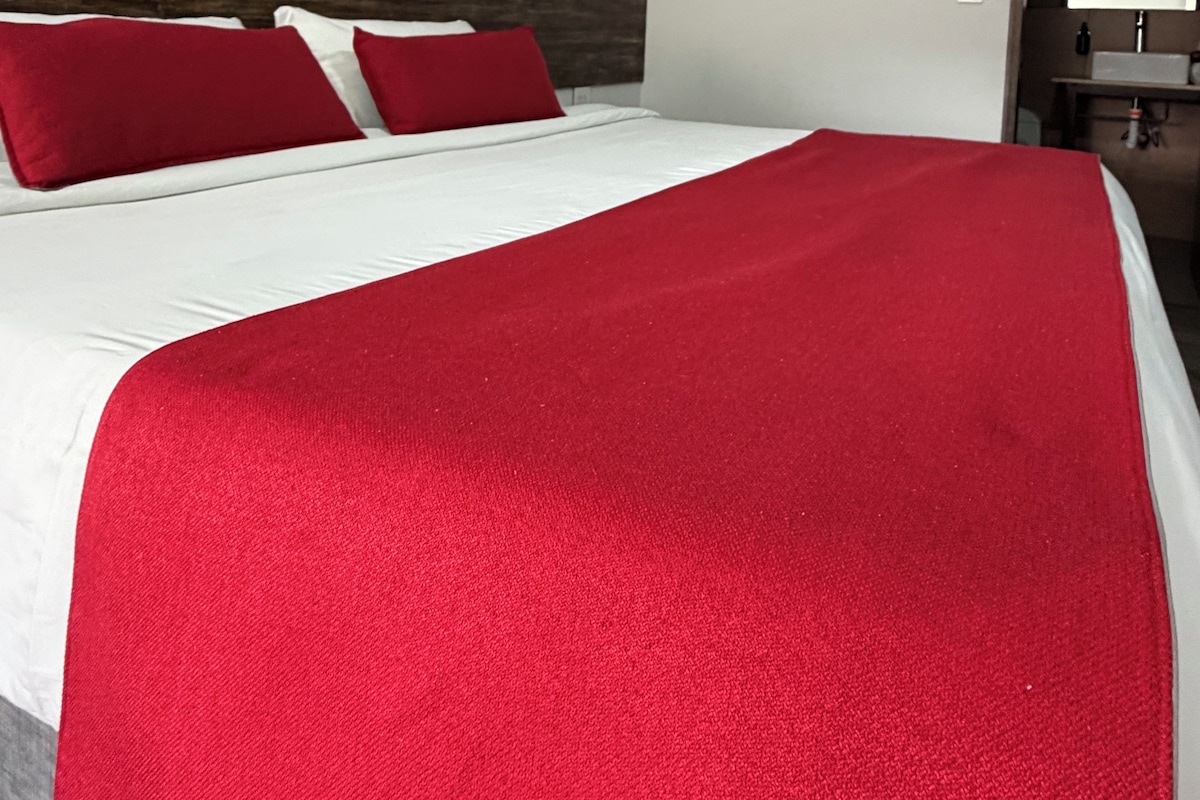 A hotel room with a bed, red pillows, a red throw blanket, a wooden headboard, and a mirror.