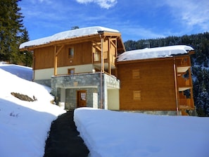 A two-story wooden chalet with a stone foundation, surrounded by snow-covered ground and trees. - Apartment with south-facing terrace - 4 guests (Crest-Voland)