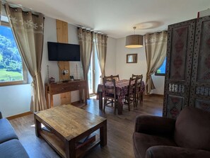 A room with a wooden dining table, chairs, a television on the wall, and a large window with curtains. - Cozy apartment with Mont-Blanc view - 6 guests (Hauteluce)