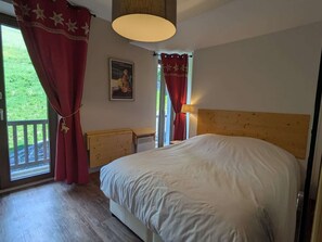 A bedroom with a large bed, a wooden headboard, a nightstand, a lamp, and a window with red curtains. - Cozy apartment with Mont-Blanc view - 6 guests (Hauteluce)
