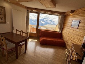 A cozy room with a wooden floor, a dining table with chairs, a sofa, and a large window with a mountain view. - Cozy apartment in Alpe d'Huez - 5 guests (Huez)